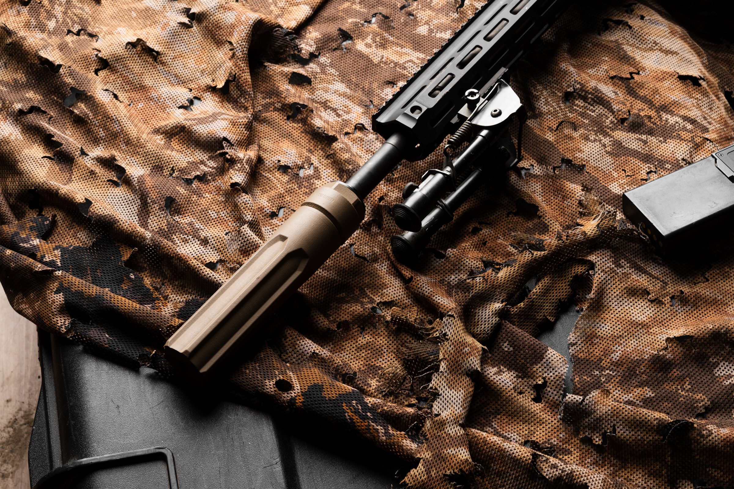 FDE Overwatch suppressor at the muzzle end of a precision rifle laid on ghillie camouflage netting