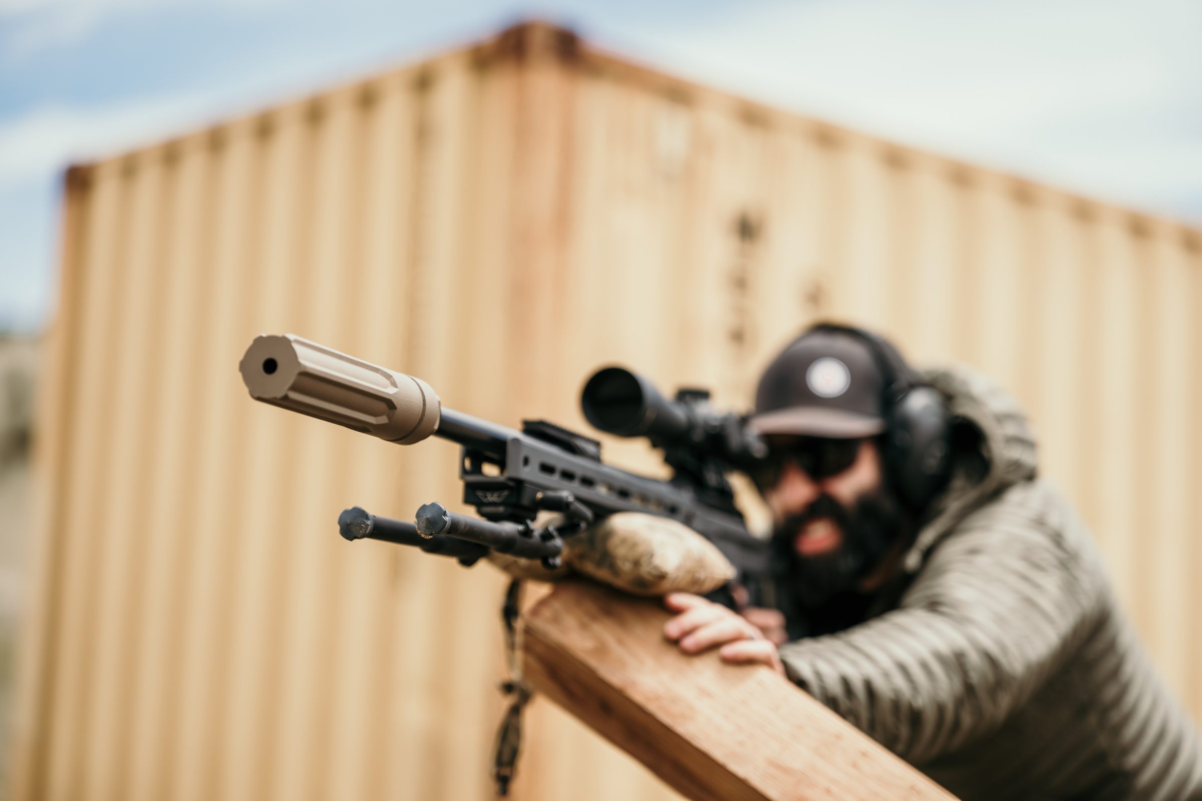 Operator in ballistic mask over wooden barrier, precision rifle with Overwatch pointed downrange