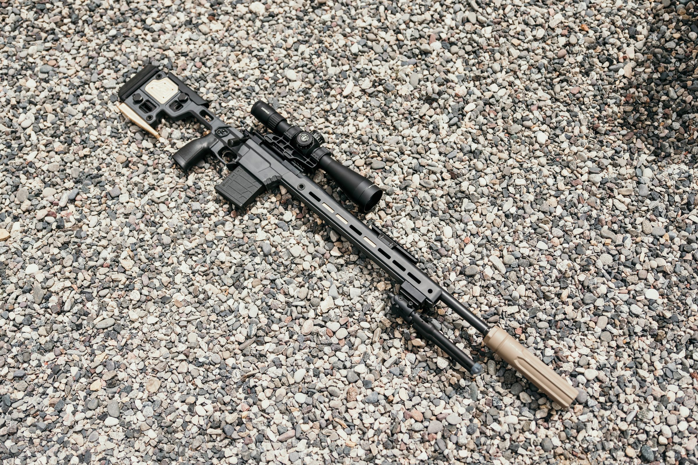 Precision rifle with Overwatch suppressor laid flat on gravel, low-angle documentary composition