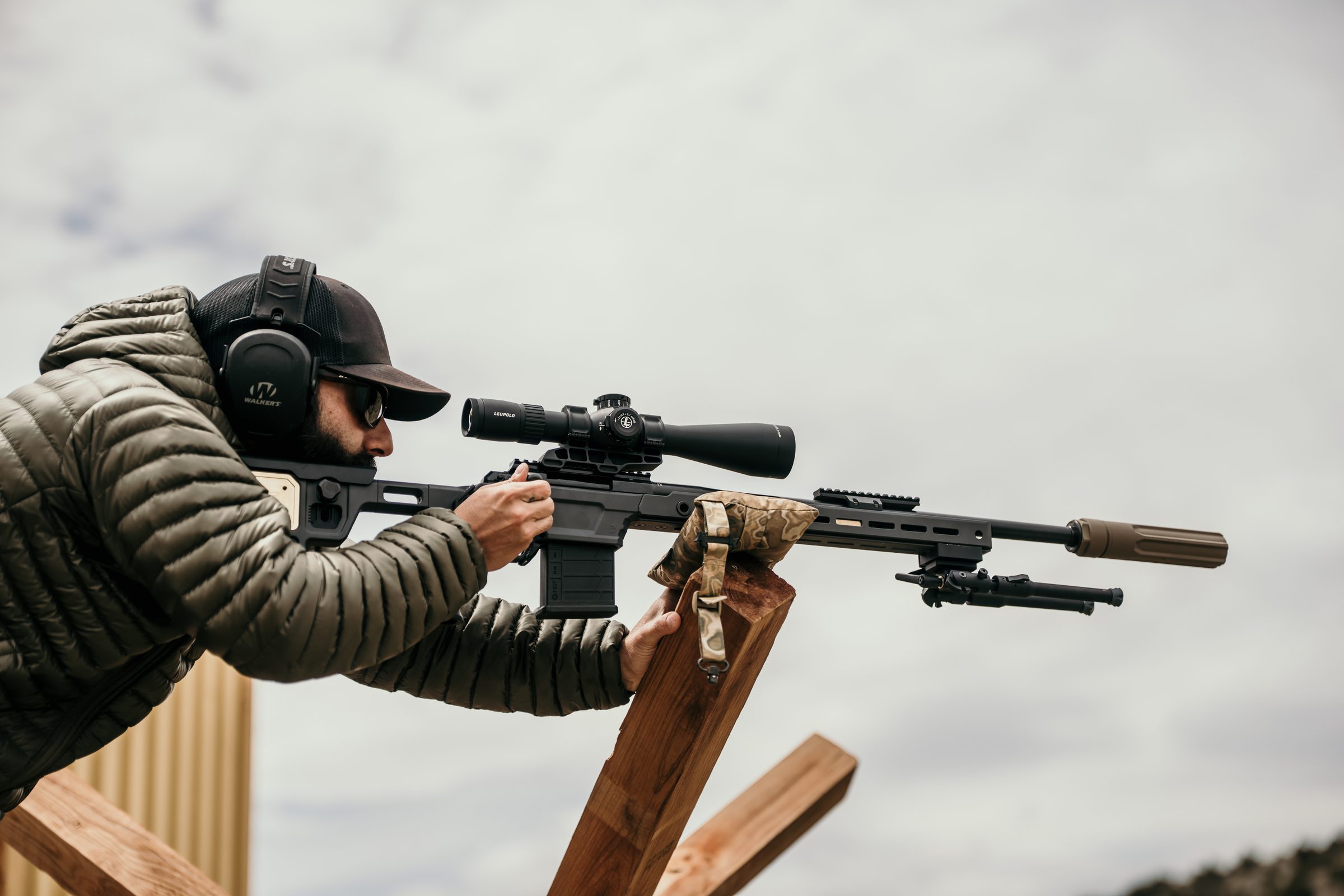 Shooter in olive jacket shouldering an FDE-Overwatch-suppressed precision rifle over a wooden barrier